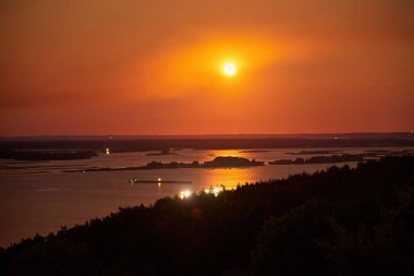 Gece ay ışığında ışıldayan bir nehrin büyüleyici görüntüsü sakin sulara sıcak bir yansıma yapıyor. Siluetli adalar ve uzak ışıklar dingin ve büyülü atmosfere eklenir.