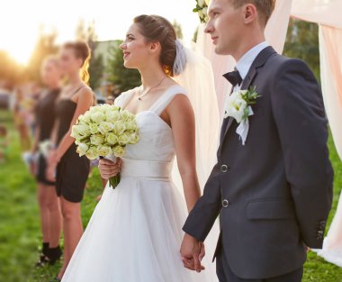 Beyaz bir gül buketi tutan bir gelinin ve düğün törenlerinde damadın elini tutarken zarif gelinlik ve damadın elbisesini yaka çiçeğiyle renklendirirken..