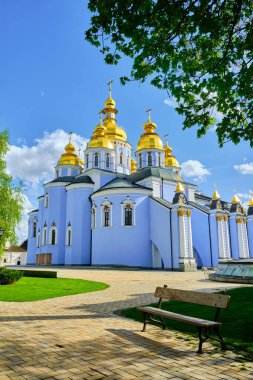 Ukrayna 'nın başkenti Kyiv' deki St. Michael 's Golden Domed Manastırı, Mykhailivska Meydanı' ndan izleniyor. Ön planda ahşap bir bank var ve manastırın mavi cephesi ve altın kubbeleri ön planda..