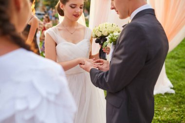 Damat tören sırasında gelinin parmağına bir alyans takar. Gelin elinde bir buket beyaz gül tutarken ikisi de süslü bir kemerin altında akan kumaşla yemin ediyor..