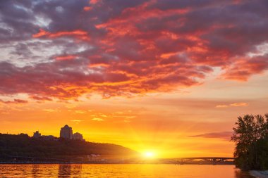 Ukrayna 'nın Kyiv kentindeki Dnipro Nehri üzerinde yoğun ve renkli bir gün batımı. Önünde kumlu bir plaj var. Gökyüzü turuncu, kırmızı ve morun karışımıdır, suyu yansıtır..