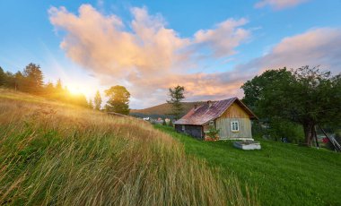 Dramatik bir gökyüzünün altında çimenli bir dağ yamacındaki kırsal ahşap bir evin manzarası. Bulutlar renkli günbatımı ışığını yakalıyor. Kırsal manzara manzarası.
