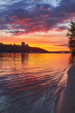 Ukrayna, Kyiv 'deki Dnipro Nehri üzerinde canlı bir günbatımı. Su, ateşli turuncu ve kırmızı gökyüzünü yansıtıyor. Şehir silueti uzaklarda..