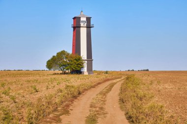 Uzun, çizgili bir deniz feneri (kırmızı, beyaz ve muhtemelen siyah renkli bantlar) açık, açık ve kuru bir alanda açık mavi bir gökyüzünün altında duruyor. Yanında tek bir ağaç var..