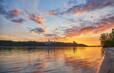 Gün batımında Ukrayna, Kyiv 'in panoramik manzarası. Dnipro Nehri renkli gökyüzünü yansıtır, şehir manzarası ve Anavatan Anıtı uzaklardan görülebilir.