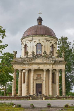 Klasik mimarinin bulunduğu tarihi bir kilisenin ön cephesinde sütunlar, heykeller ve bulutlu gökyüzü altındaki ağaçlar tarafından çerçevelenmiş önemli bir kubbe yer alıyor..