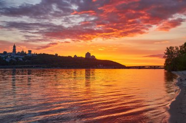 Ukrayna, Kyiv 'deki Dnipro Nehri üzerinde canlı bir günbatımı. Su, ateşli turuncu ve kırmızı gökyüzünü yansıtıyor. Şehir silueti uzaklarda..
