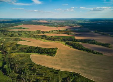 Yüksek hava manzarası geniş bir tarımsal manzarayı gösteriyor. Kahverengi tarlalar ve yeşil ormanlardan oluşan bir mozaik bulutlu mavi gökyüzünün altında ufka doğru uzanıyor..