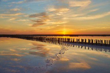 Pembe ve turuncu bulutlu pastel gün batımı ufukta eski ahşap yapılar ve pembe tuz gölünün sakin sularında yansıyor..