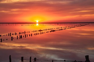 Sakin bir tuz gölünün üzerinde nefes kesici bir gün batımı. Kırmızı, turuncu ve pembe bulutlu canlı, ateşli gökyüzü durgun suda mükemmel bir şekilde yansıyor. Eski tahta direkler sıraya dizilir.