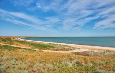 Toprak bir yol çimenli bir tarladan geçerek parlak mavi bir gökyüzünün altında ince bulutlu bir göle çıkar. Yaz manzarası, seyahat ve keşif konsepti.
