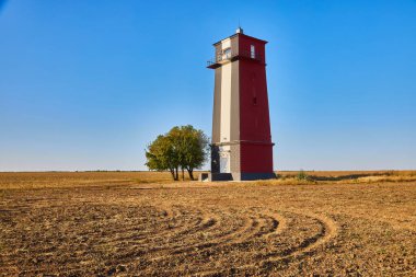 Uzun, çizgili bir deniz feneri (kırmızı, beyaz ve muhtemelen siyah renkli bantlar) açık, açık ve kuru bir alanda açık mavi bir gökyüzünün altında duruyor. Yanında tek bir ağaç var..