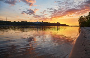 Ukrayna, Kyiv 'deki Dnipro Nehri üzerinde renkli bir günbatımı. Şehrin ufuk çizgisi, tarihi binalar da dahil, ön planda kumlu bir plaj ile uzaktan görülebilir..