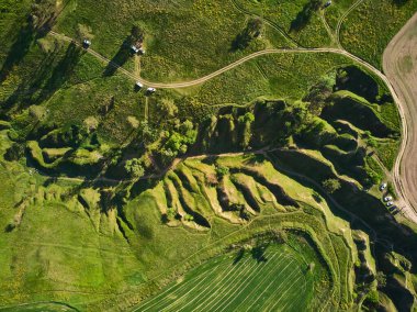 Hava aracı perspektifi aşınmayı gösteren yuvarlanan yeşil tepeler ve aşağıda park edilmiş arabalar. Arkaplanda mavi bulutlu gökyüzünün altında düz tarım alanları yer alıyor.