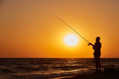 Canlı bir günbatımında sahilde oltasıyla güçlü bir alçı yapan bir balıkçının silueti..