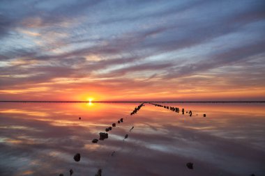 Sakin bir tuz gölünün üzerinde nefes kesici bir gün batımı. Kırmızı, turuncu ve pembe bulutlu canlı, ateşli gökyüzü durgun suda mükemmel bir şekilde yansıyor. Eski tahta direkler sıraya dizilir.