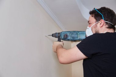 Emniyet gözlüğü takan ve maske takan kişi tavana yakın bir duvara delik açıyor. Ev içi geliştirme, DIY ya da inşaat işine yakın çekim.