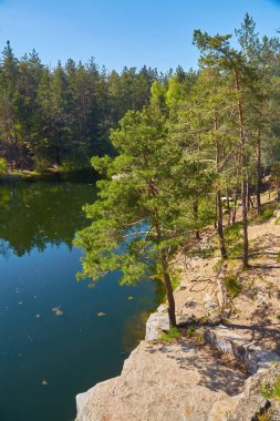 Sel basmış granit ocağının geniş manzara manzarası. Dik kayalıklar ve yemyeşil orman berrak güneşli gökyüzünün altındaki derin, sakin mavi suya yansıyor..