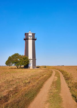 Khablovsky Deniz feneri (muhtemelen), uzun, çizgili bir deniz feneri, kırsal, çimenli bir arazide duran ve ona doğru giden toprak bir yol. Açık mavi gökyüzü.