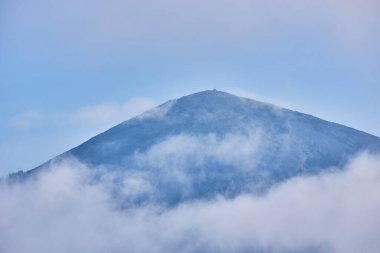 Soluk bir gökyüzüne doğru dönen sisle kısmen gizlenmiş bir dağın zirvesinin yakın görüntüsü. Hava durumunu ve ıssızlığı vurgulayan atmosferik sahne.