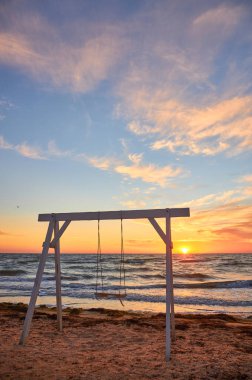 Güneş ufukta batarken, kumlu bir sahilde beyaz ahşap bir salıncak duruyor. Denizde ve gökyüzünde, dağınık bulutlarla ılık bir altın parıltı yayıyor..