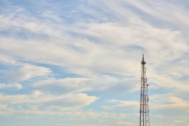Uzun bir hücresel kule, ince beyaz bulutlarla dolu mavi bir gökyüzüne karşı duruyor. Haberleşme ve teknoloji kavramı.
