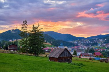 Köyün ahşap ahırı vadideki bir dağ köyüne tepeden bakan çimenli bir yamaçta duruyor. Dramatik, renkli günbatımı bulutları gökyüzünü dolduruyor. Atmosferik görünüm.