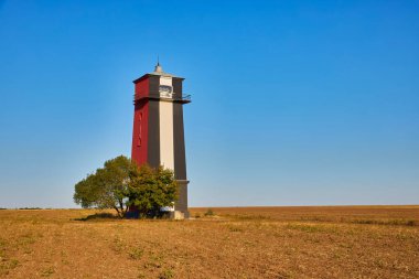 Uzun, çizgili bir deniz feneri (kırmızı, beyaz ve muhtemelen siyah renkli bantlar) açık, açık ve kuru bir alanda açık mavi bir gökyüzünün altında duruyor. Yanında tek bir ağaç var..