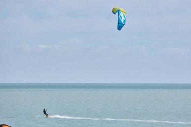 Birkaç uçurtmacı sakin, turkuaz bir denizde güneşli bir günün tadını çıkarıyorlar. Uçurtmalar havada yüksekte, bir uçurtmacı suda görünür..