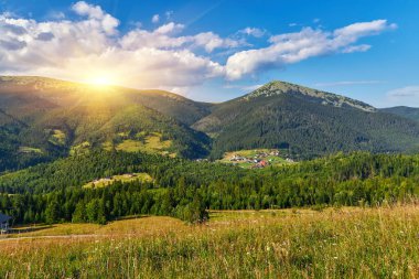 Panoramik yaz manzarası. Dağ köyüne bakan yeşil ormanlık tepelerin arasına kurulmuş. Parlak mavi gökyüzünün altında dramatik beyaz kümülüs bulutları..