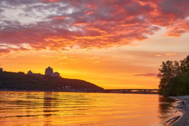 Ukrayna 'nın Kyiv kentindeki Dnipro Nehri üzerinde yoğun ve renkli bir gün batımı. Önünde kumlu bir plaj var. Gökyüzü turuncu, kırmızı ve morun karışımıdır, suyu yansıtır..