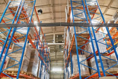 View looking up between towering blue and orange pallet racks in a warehouse. The high shelving is heavily stocked with shrink-wrapped goods. High-density industrial storage.