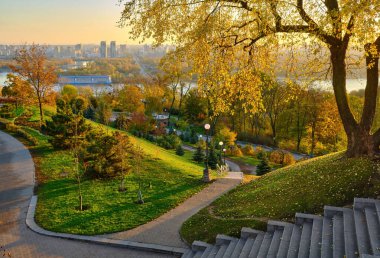 A scenic panoramic view of the Dnipro River and the left bank of Kyiv from a hilltop park at sunrise. The warm light illuminates the vibrant yellow autumn foliage on the trees.