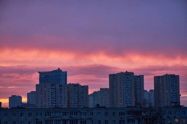 Siluetli bir Kyiv şehri silueti pembe, turuncu ve mor bulutlu canlı bir günbatımı gökyüzüne karşı görünür. İnşaat vinçleri binaların arasında duruyor ve sahne sakin suya yansıyor..