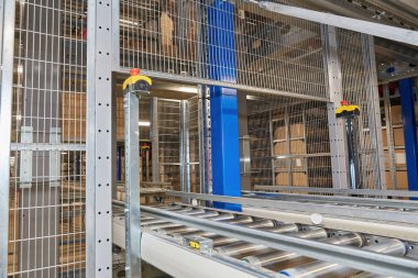 Interior of a modern warehouse or distribution center featuring an extensive automated conveyor belt system for package sorting. Shelves stacked with goods and boxes in background.