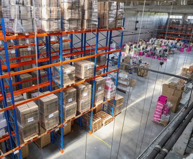 High angle overview of a spacious and busy warehouse, showcasing tall pallet racks heavily stocked with goods, alongside conveyor systems and stacks of boxes