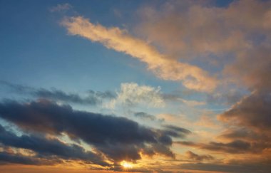 Birden fazla bulut katmanıyla dolu dramatik bir günbatımı gökyüzü. Koyu gri bulutlar ince bulutların altında süzülür. Ufukta parıldayan güneşin altın ışığını yakalar..
