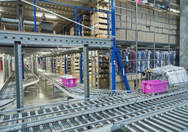 Interior of a modern warehouse or distribution center featuring an extensive automated conveyor belt system for package sorting. Shelves stacked with goods and boxes in background.