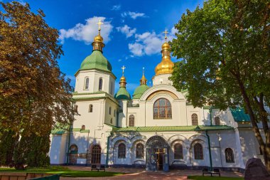 View of Saint Sophia Cathedral in Kyiv, its iconic green and golden domes peeking through lush green trees on a sunny day. A serene perspective of the historic site.