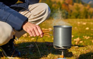 Bir adamın eli, dağların arka planında yemek yemek için bir tencere tutar, bir fast food sistemi, güvenli bir gaz ocağı, yürüyüş ekipmanları, yürüyüş aletleri..