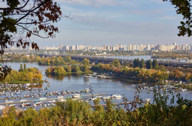 Panoramic autumn view of Kyiv and the Dnipro River from a high vantage point, showing a marina full of boats, a bridge, and the city skyline. Trees show vibrant fall colors.