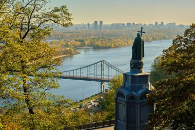 A scenic autumn view from Volodymyrska Hill in Kyiv, with the Monument to Volodymyr the Great in the foreground overlooking the Parkovy Pedestrian Bridge, the Dnipro River, and the distant city skyline at sunrise.