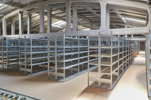 Interior of a large, empty warehouse featuring rows of metal shelving units under an arched concrete ceiling with skylights. A roller conveyor system is in the foreground.
