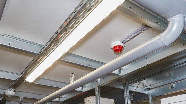 Close-up low-angle view of an industrial ceiling in a warehouse, showing a fire sprinkler head, red fire alarm, and fluorescent lighting fixture. Safety infrastructure.