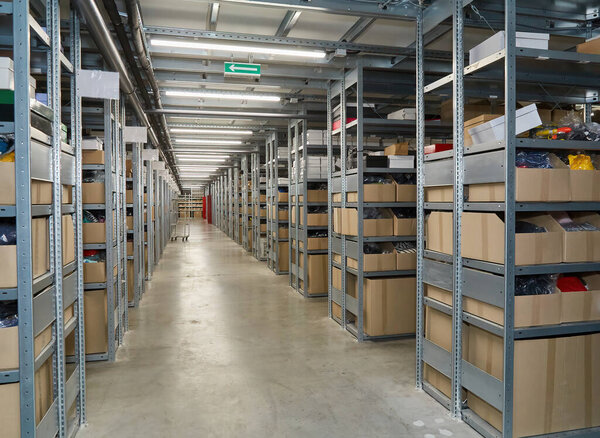 View down a long, well-lit aisle in a warehouse, lined with tall metal shelving units neatly stocked with cardboard boxes and other inventory. Organized storage facility.