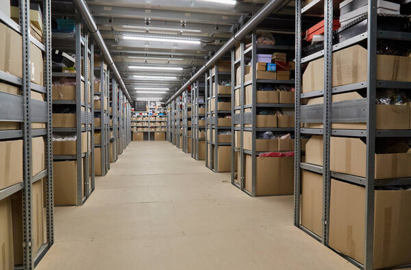 Perspective view down a long, narrow aisle in a large warehouse, flanked by towering metal shelving units densely packed with cardboard boxes. Industrial storage.