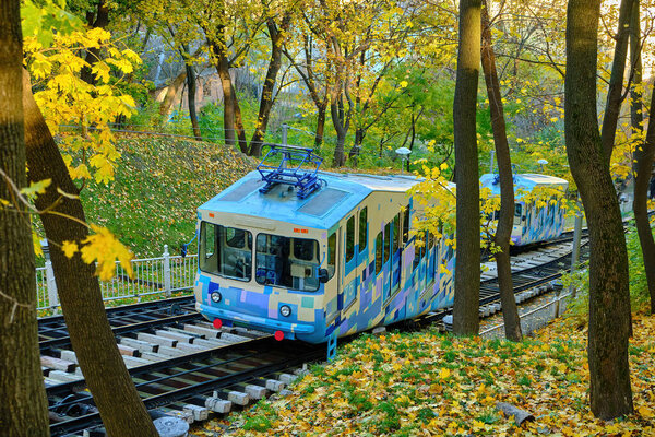 The modern car of the Kyiv Funicular ascends a steep track through a beautiful park on Volodymyr Hill, surrounded by the golden-yellow foliage of autumn.