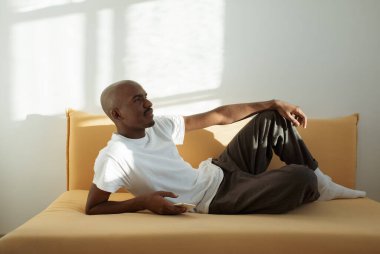 Genç siyah adam, sarı kanepede uzanmış güneş ışığıyla aydınlatılmış bir odada.