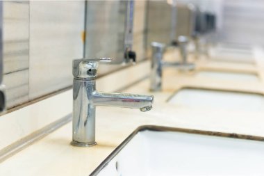 Umumi tuvaletin içi beyaz seramik lavabo, ayna ve musluklarla dolu.