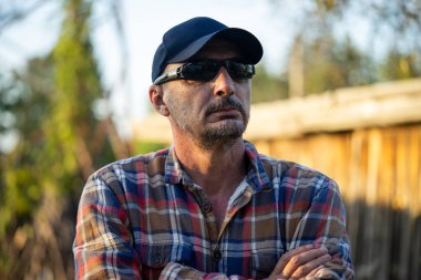 Kendinden emin, kareli gömlekli, kollarını kavuşturup köydeki kameraya bakan olgun bir adam..